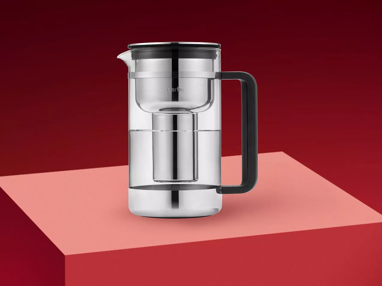 Glass water filter jug with black handle and stainless steel cartridge, on a pink block backdrop