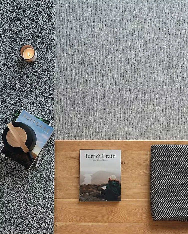 A cosy scene with a lit candle, magazines, and a book titled "Turf & Grain" on a wooden floor and textured carpet.