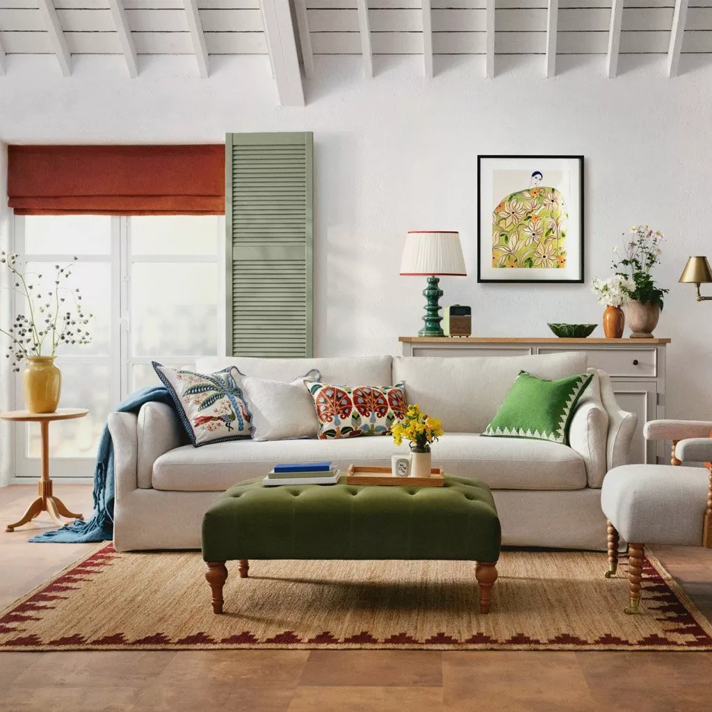 a bright and cozy living room styled with a neutral sofa, an olive green tufted ottoman, and a mix of colorful accents like patterned pillows and a sage green decorative shutter