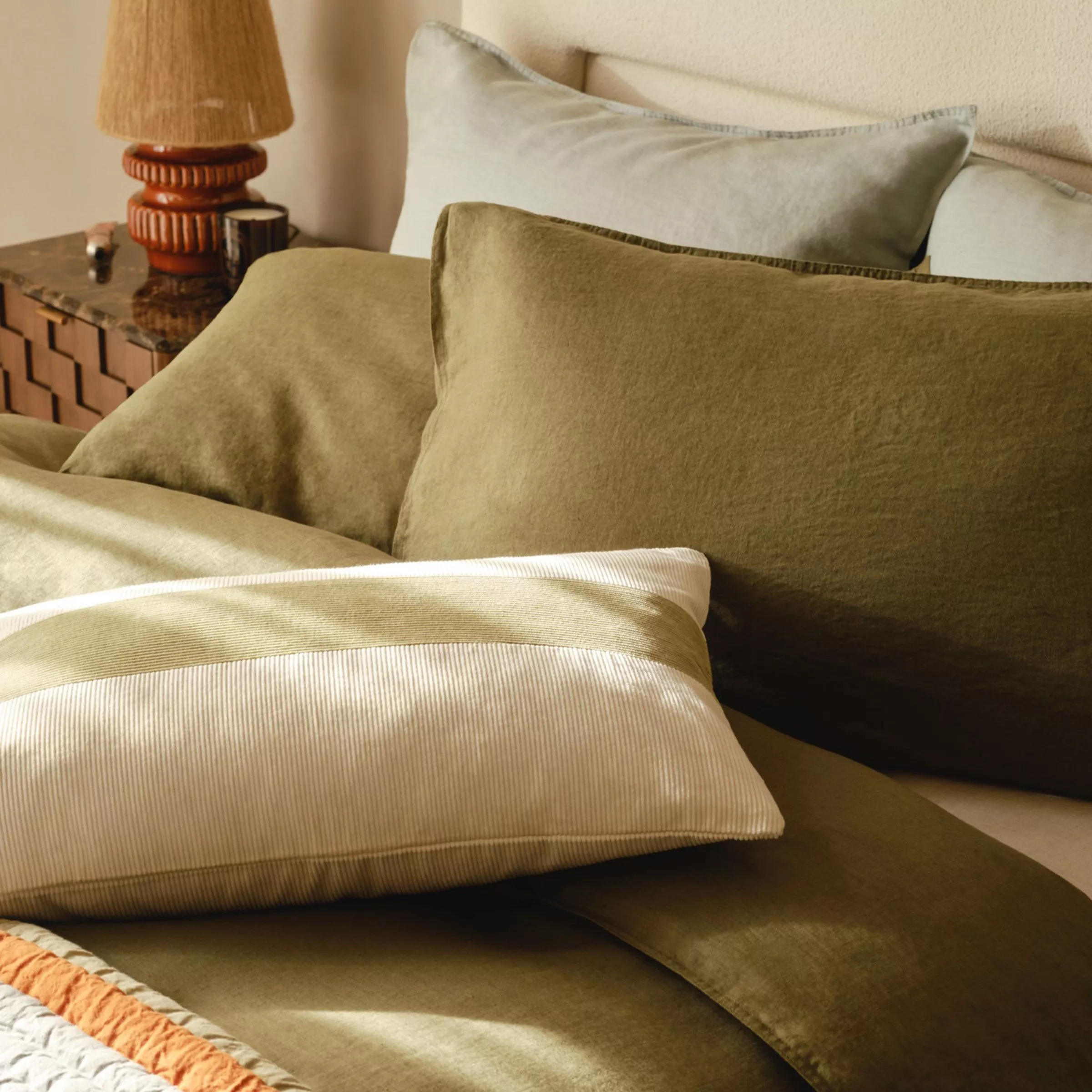 Bed made up with green linen bedding, cushion with a stripe on and bedside table was a small ratten table lamp