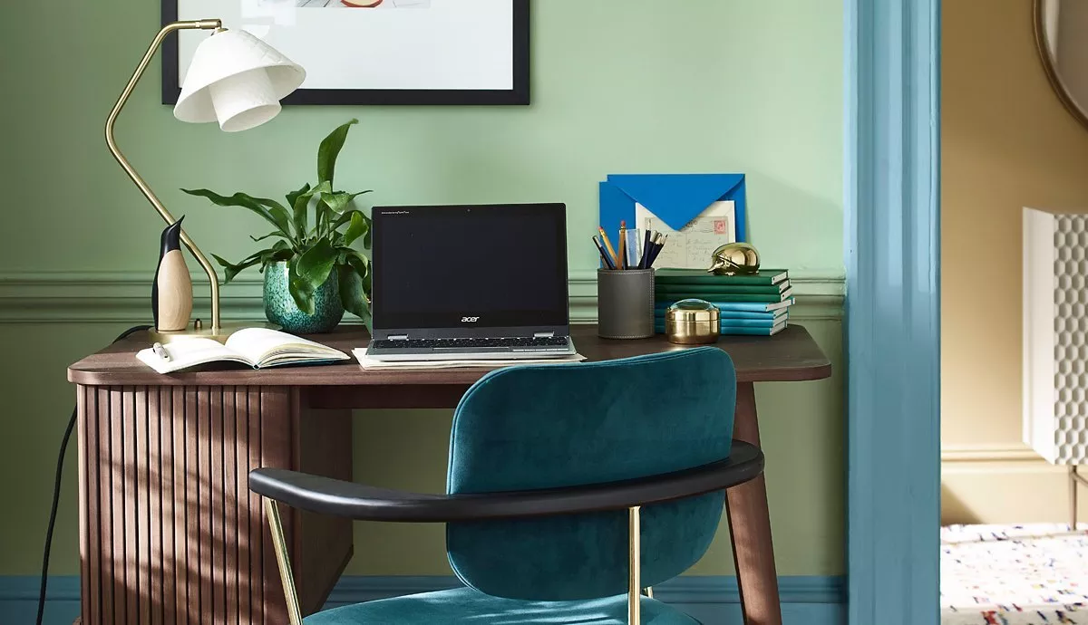 A stylish home office with a wooden desk, teal chair, and laptop. Decor includes a plant, lamp, and books.