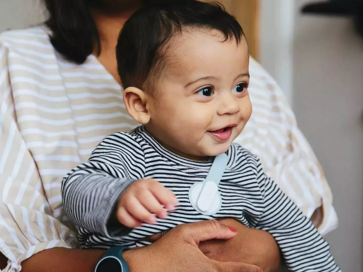 Smiling baby in striped top sitting on parent’s lap indoors, wearing clip-on baby monitor sensor on chest