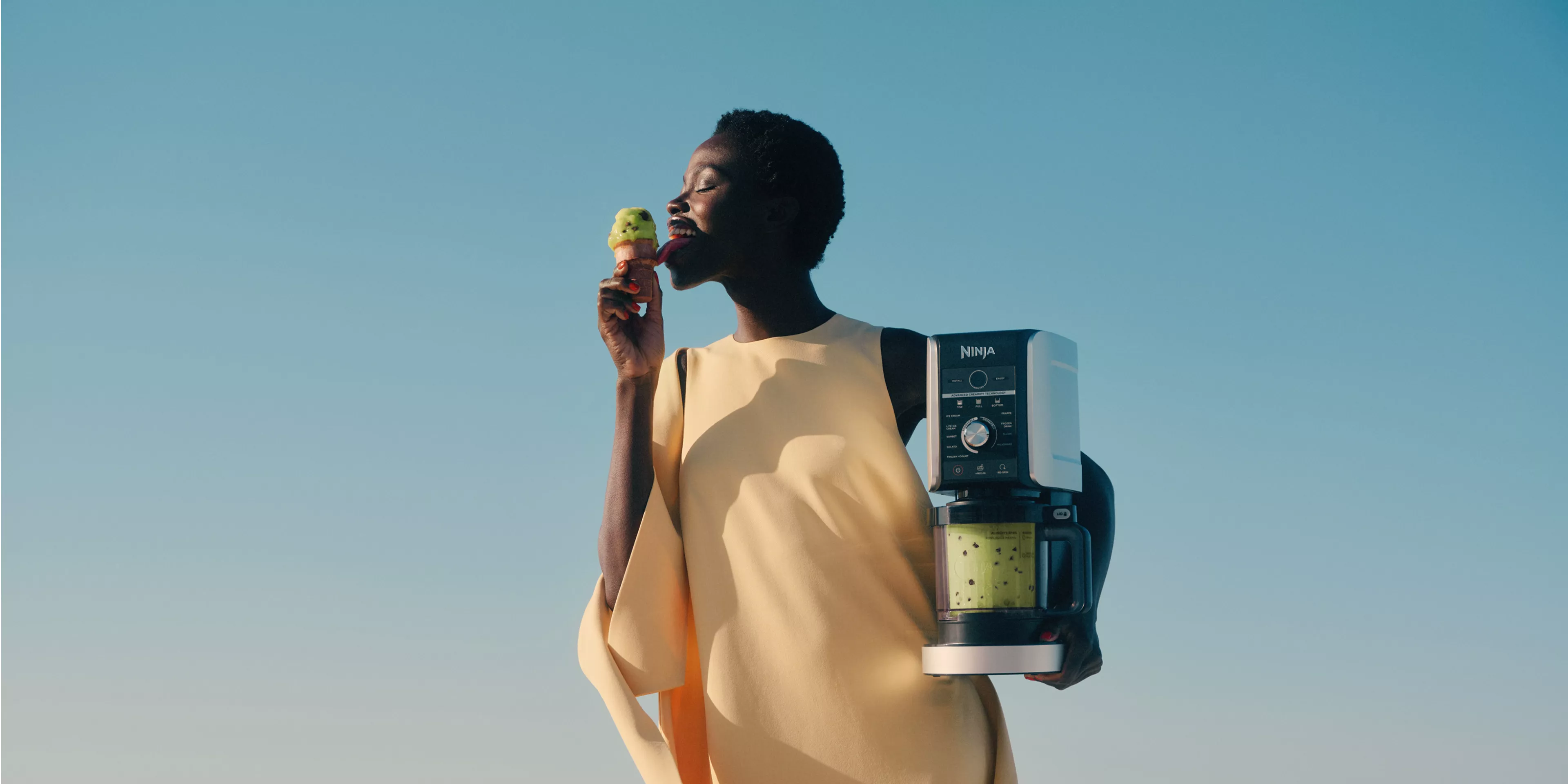 Woman eating green ice cream outdoors, holding Ninja food processor with matcha mixture against blue sky