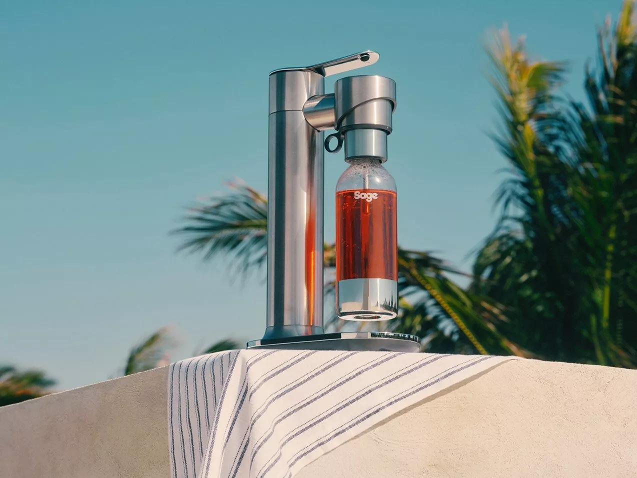 Stainless steel soda maker with bottle outdoors on terrace, palm trees behind, refreshing summer drink setup