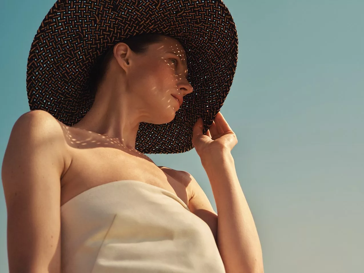 Woman in strapless dress wearing large woven sun hat, looking sideways in warm sunlight against clear blue sky