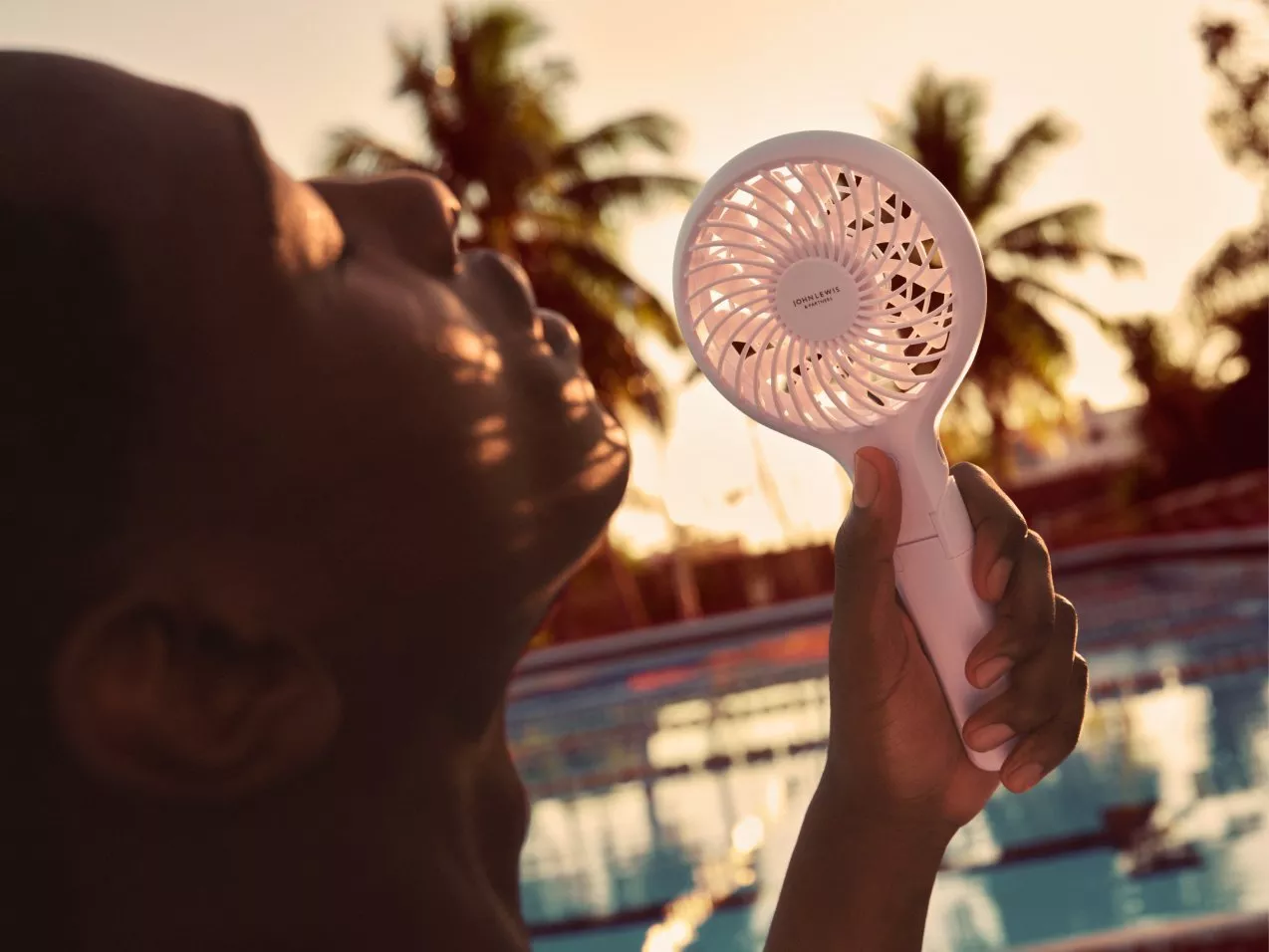 Person holding a white handheld portable fan by a swimming pool at sunset, with palm trees in the background