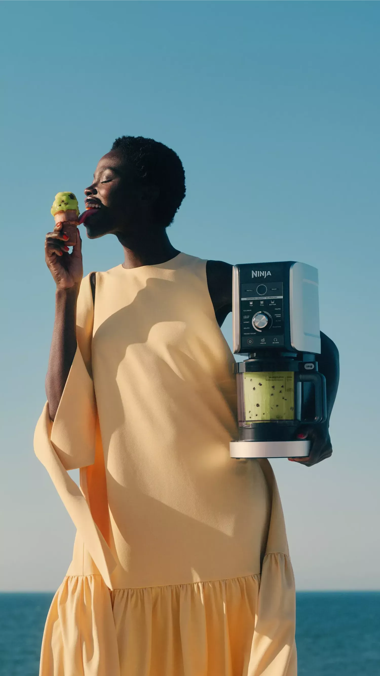 Woman eating green ice cream outdoors, holding Ninja food processor with matcha mixture against blue sky