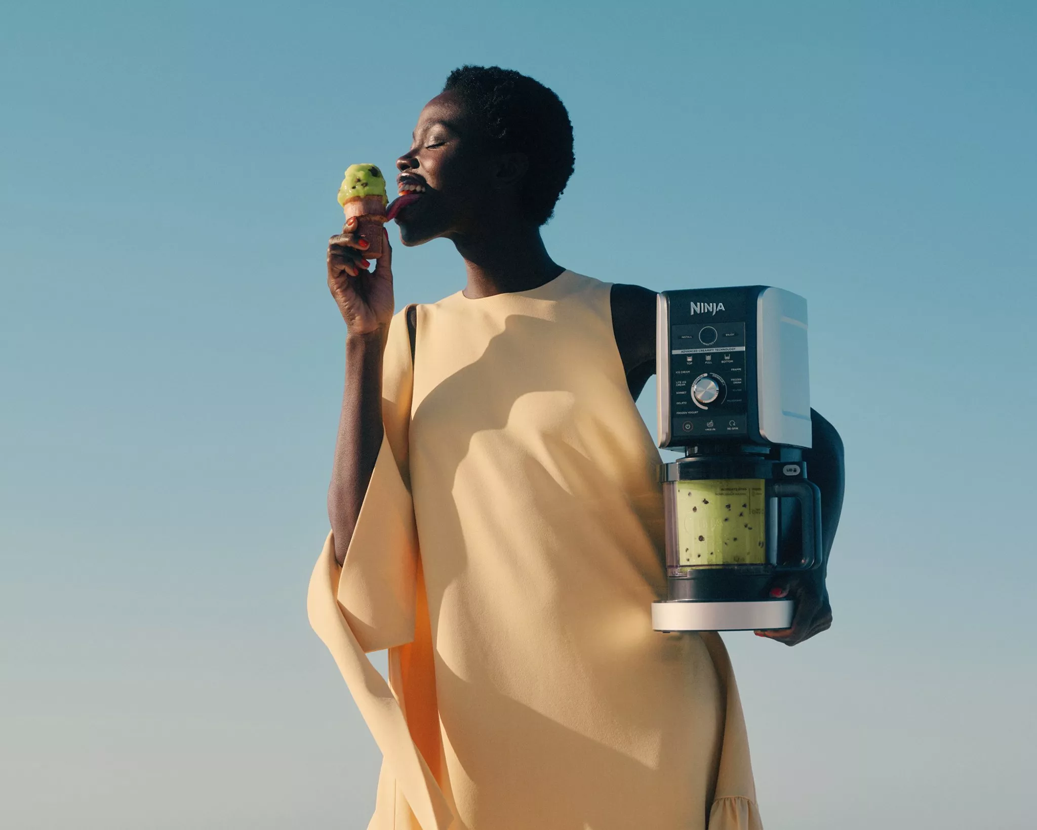 Woman enjoying ice cream while holding a Ninja Creami ice cream maker against a clear blue sky