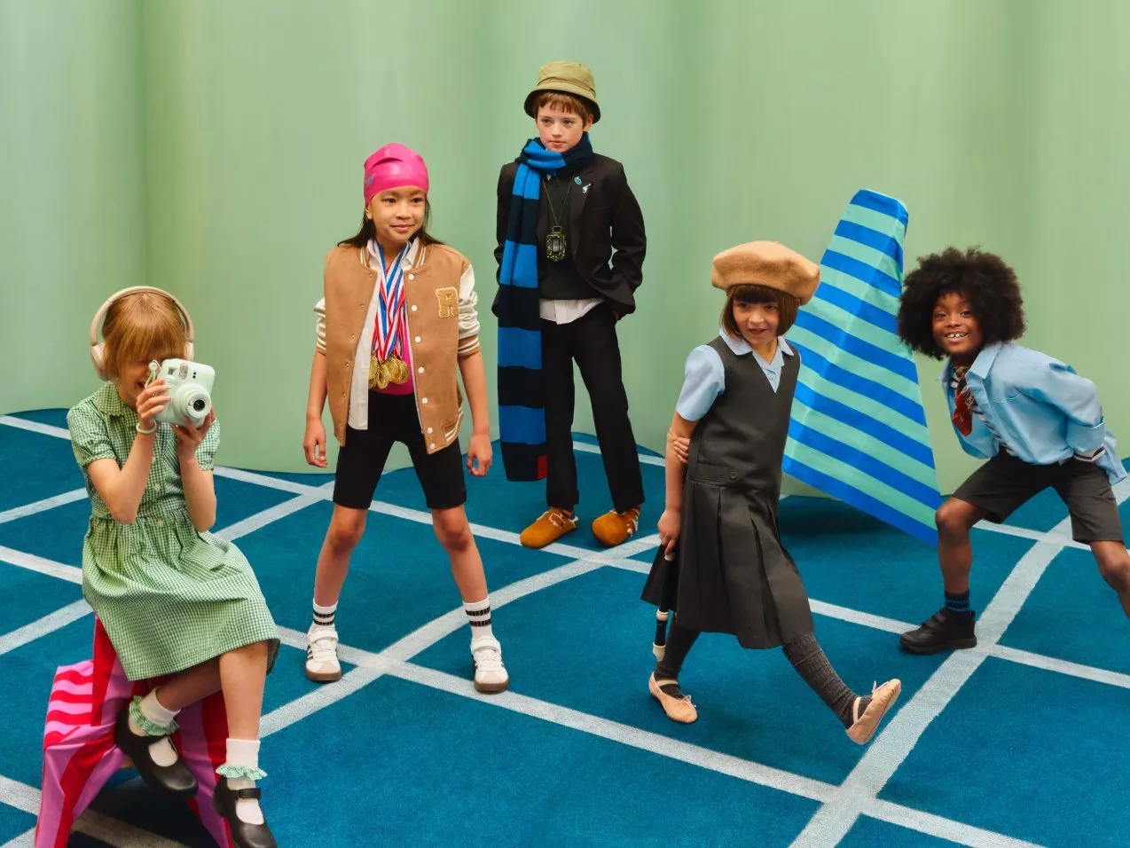 Group of children in school outfits on blue grid carpet, one taking a photo, green studio backdrop