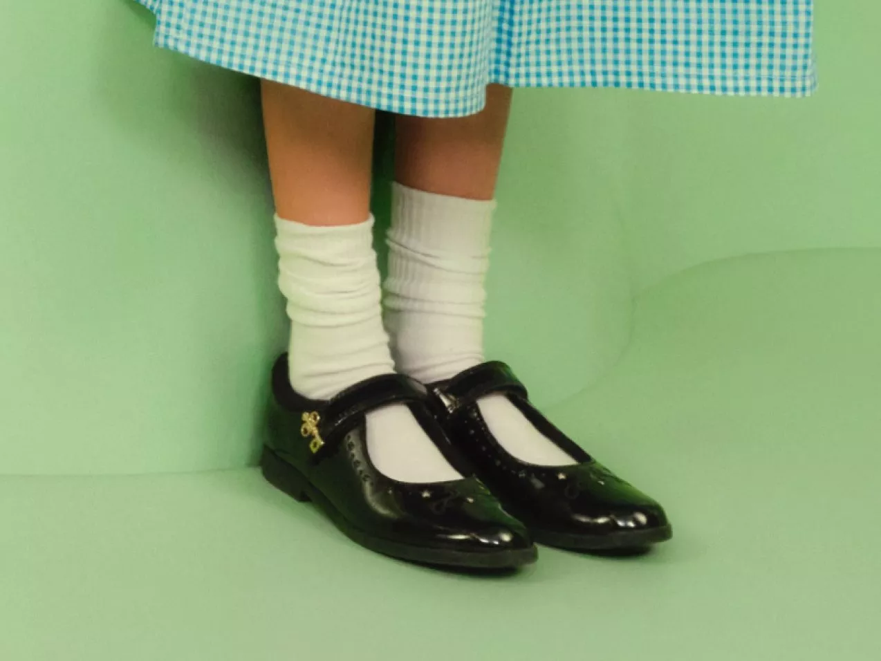 Child wearing white socks and black Mary Jane school shoes under a blue gingham dress on green backdrop