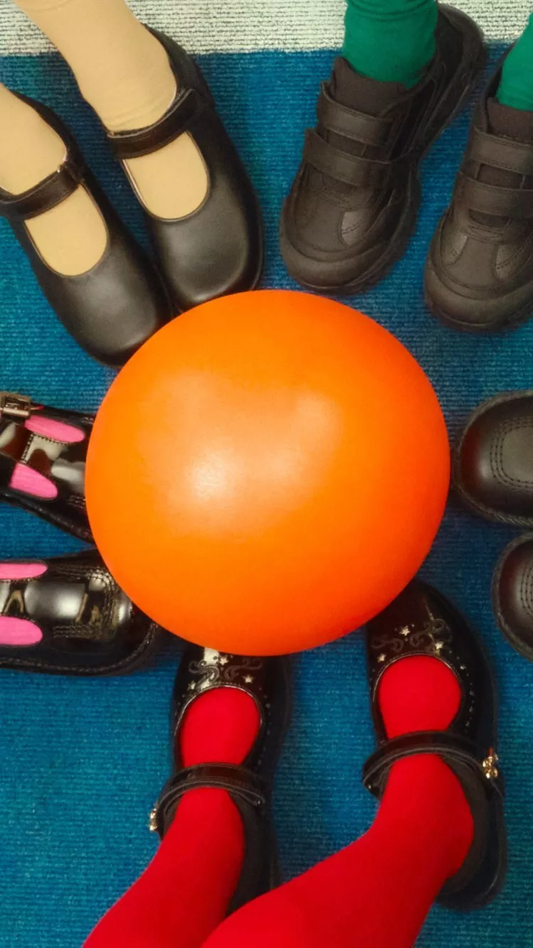 Children’s feet in school shoes around an orange ball on a blue carpet