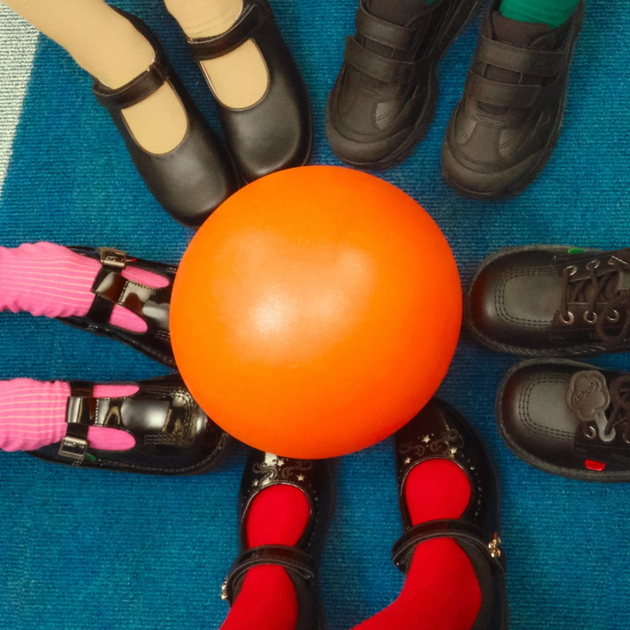 Children’s feet in school shoes around an orange ball on a blue carpet, ready for indoor playtime game