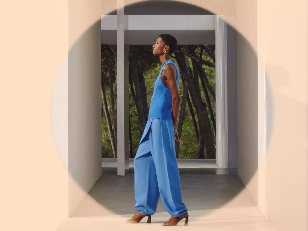 Fashion model wearing loose blue co-ord and heels in modern architectural hallway with forest view behind