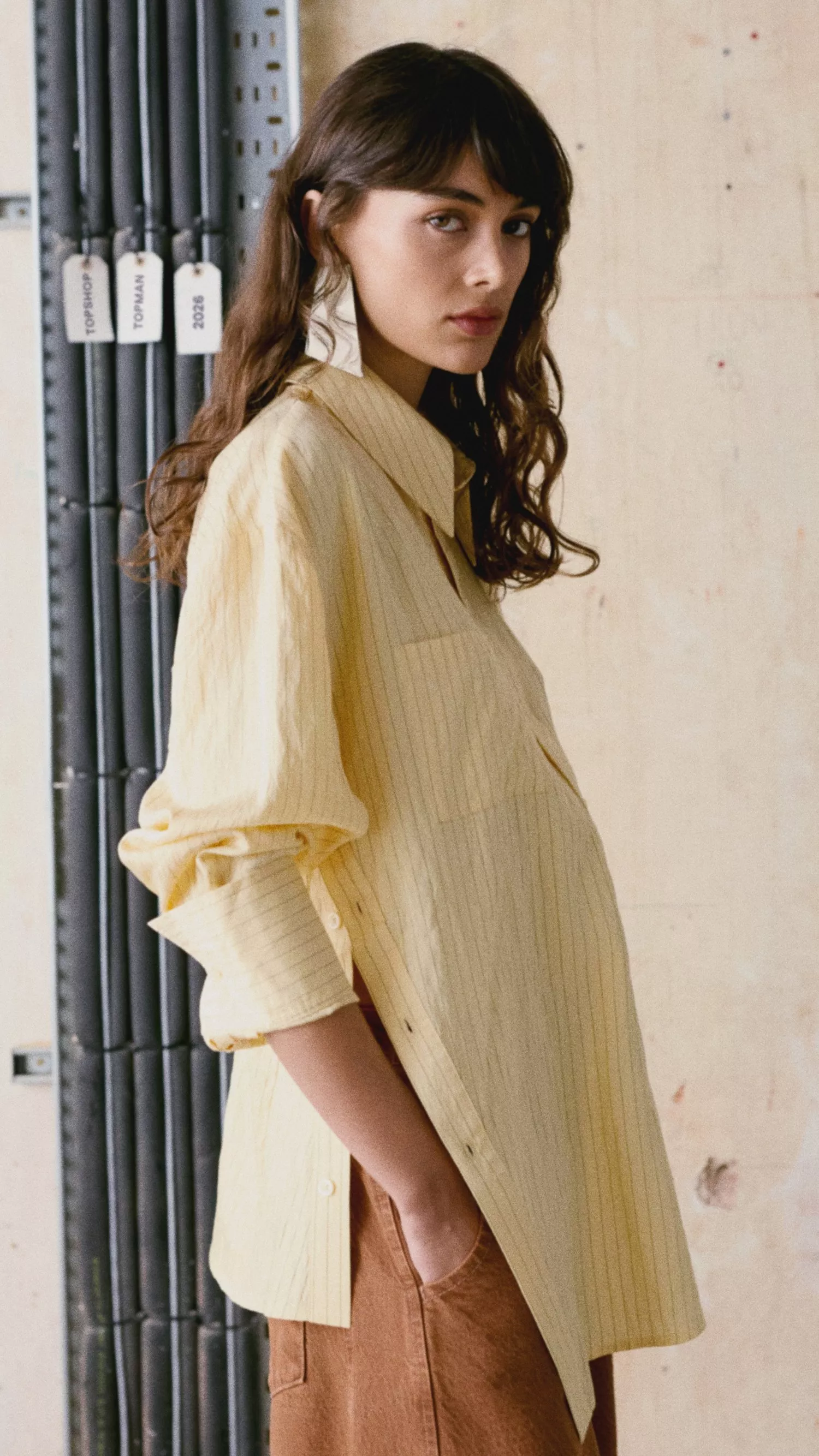 Side view of woman in oversized yellow striped shirt and brown skirt, minimalist fashion outfit against studio wall backdrop