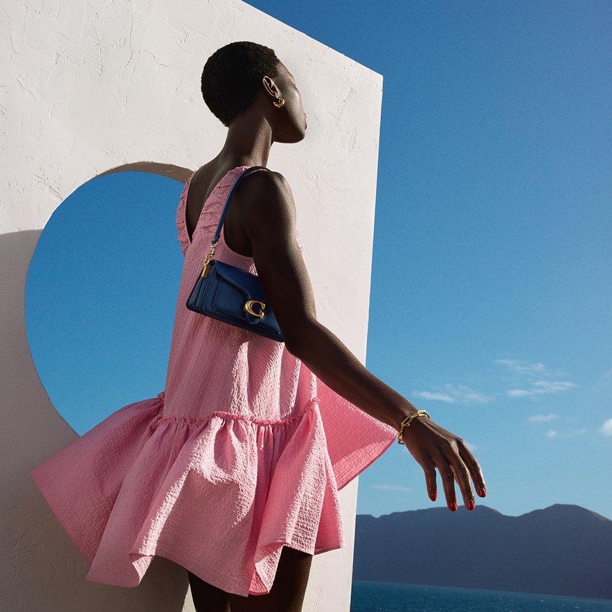 A female model wearing a pink short dress and a black Coach bag on her shoulder, outdoors.