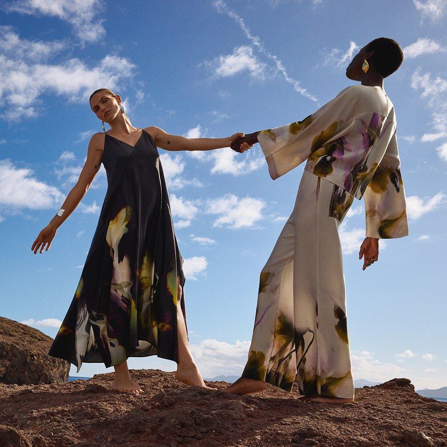 Two female models standing and holding hands — one wearing a floral dress, the other in a two-piece outfit