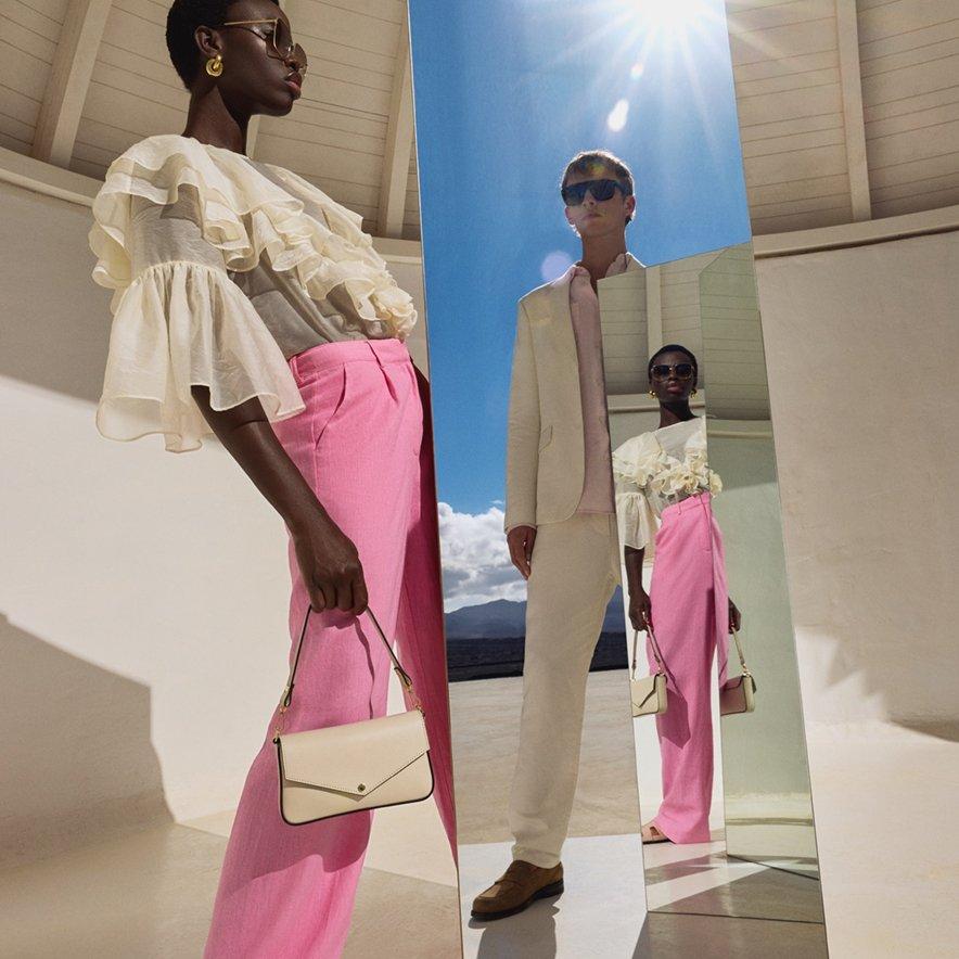 A male and a female model stand next to a mirror. One is wearing pink trousers and a shirt with large frills on the sleeves and a cream handbag, while the other is styled in a cream-coloured jacket, matching trousers