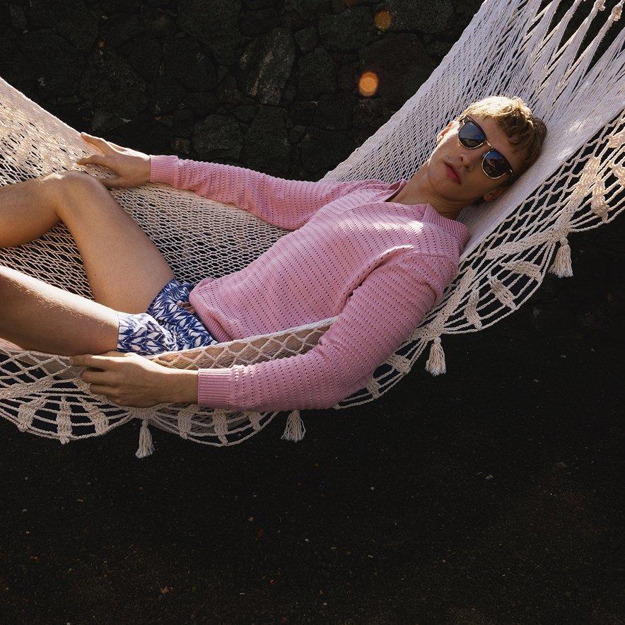 Male model wearing sunglasses, pink long-sleeve top, and blue-and-white shorts, reclining on a hammock