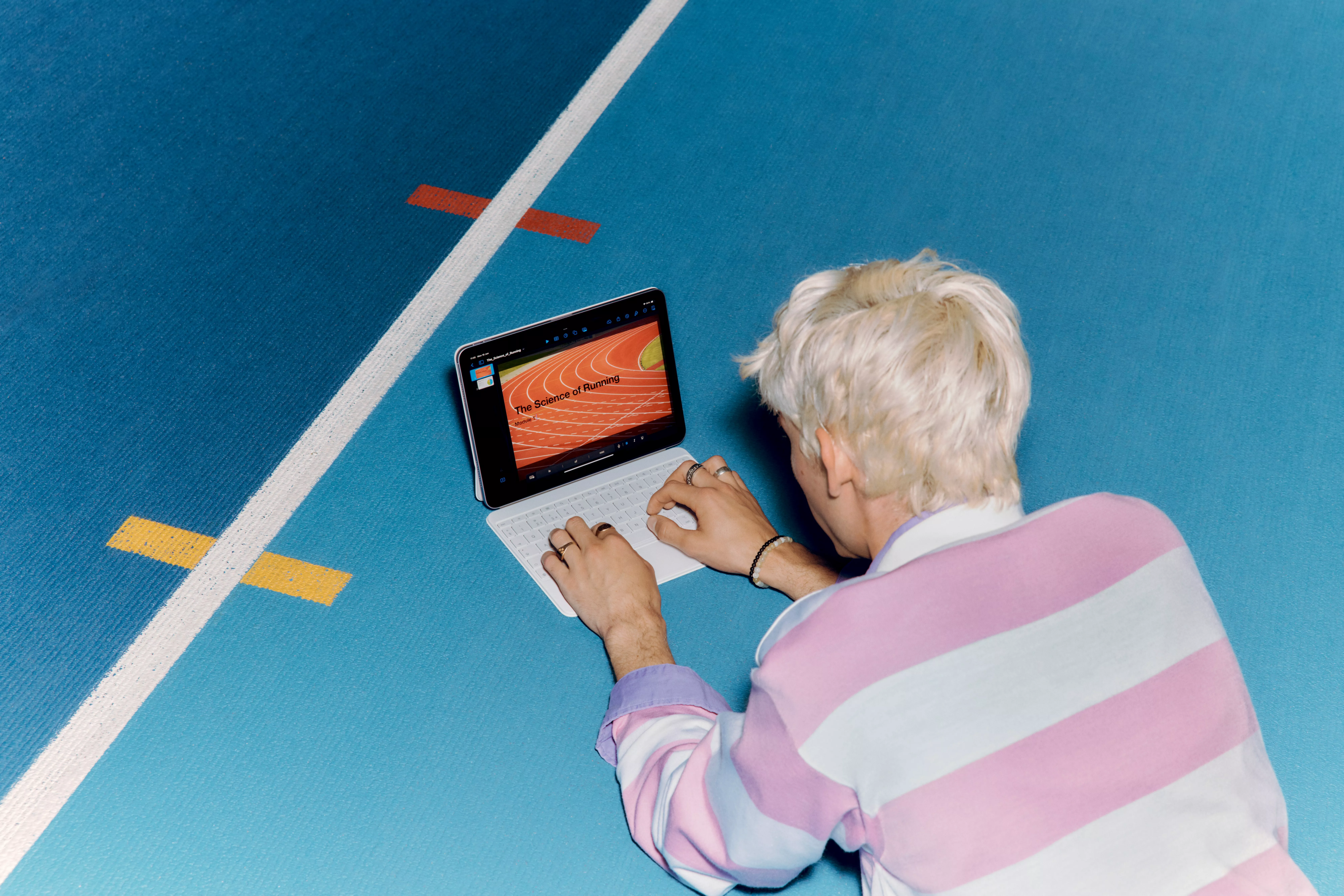 A person with blonde hair using a laptop on a blue sports court. The screen displays a presentation slide.