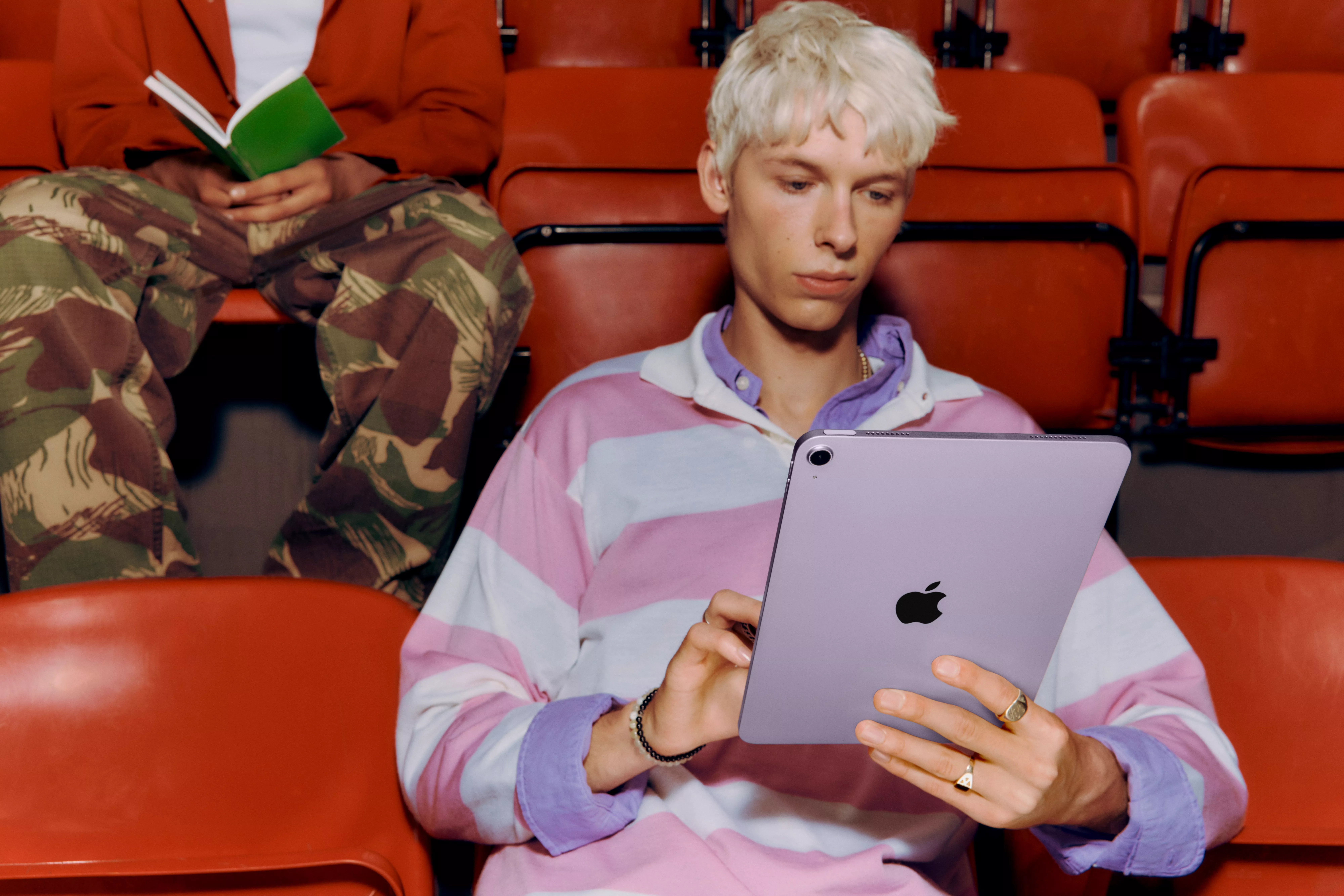 Young man using a tablet in red cinema seats, with another person reading a book in the background