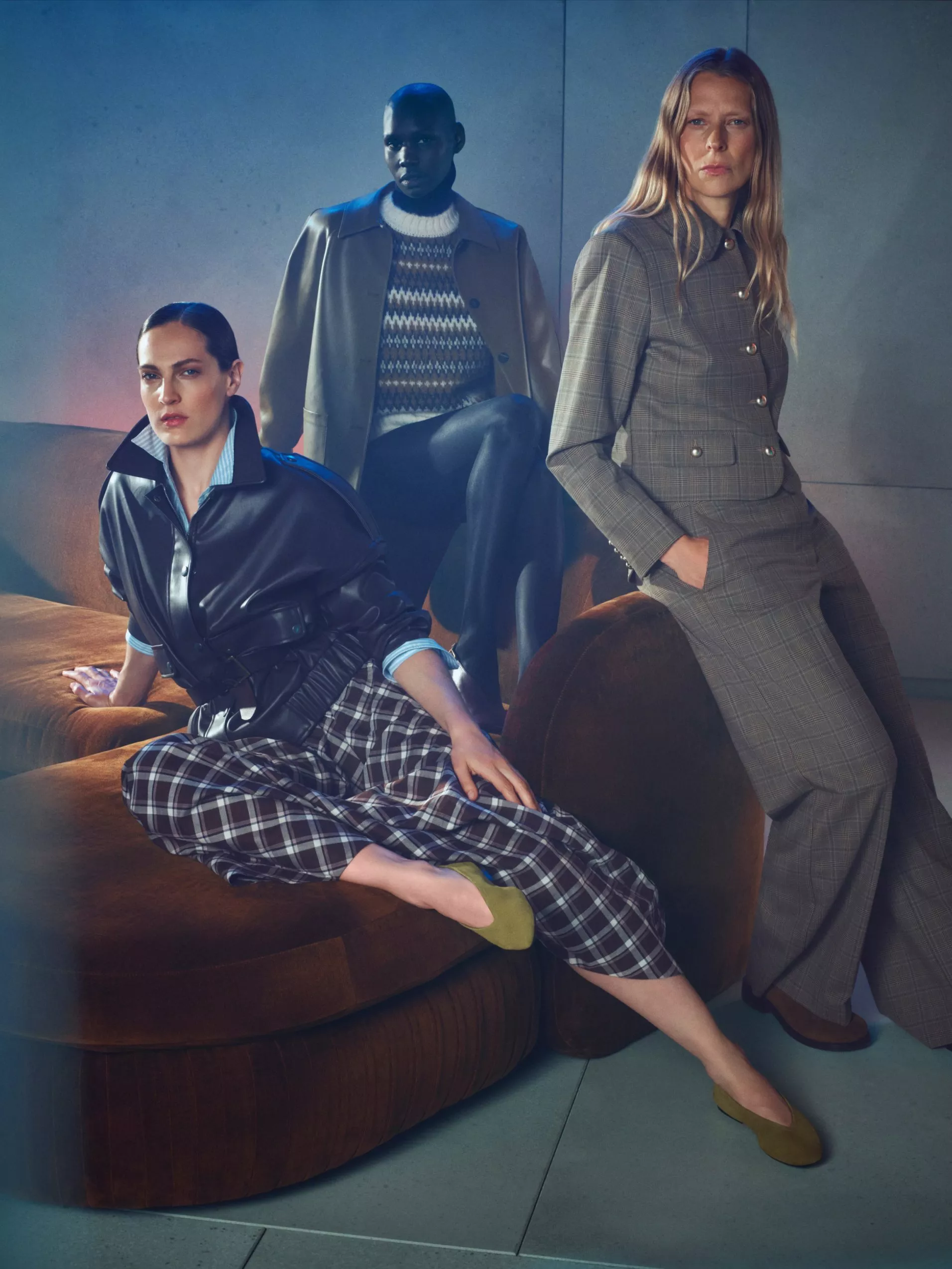 Three models posing in a dimly lit studio, wearing black leather, plaid trousers, a knit sweater, and a grey plaid suit.