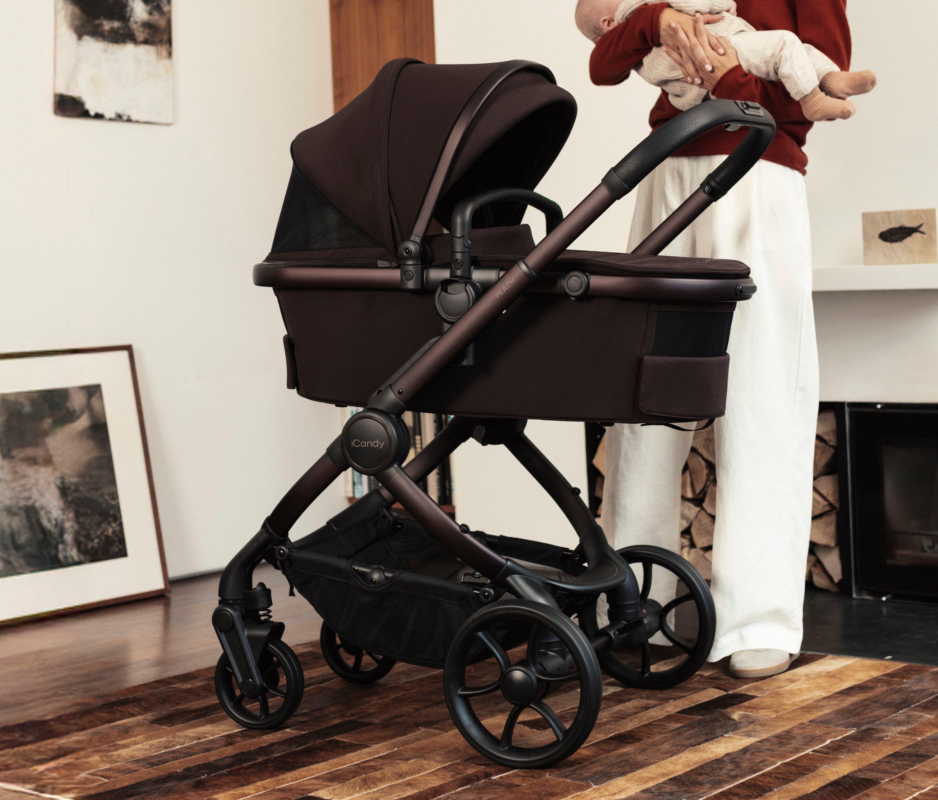 A person holds an infant next to an iCandy Peach stroller in a modern room. The dark chocolate pram features a carrycot with a large sun canopy and sleek bronze-colored framing.