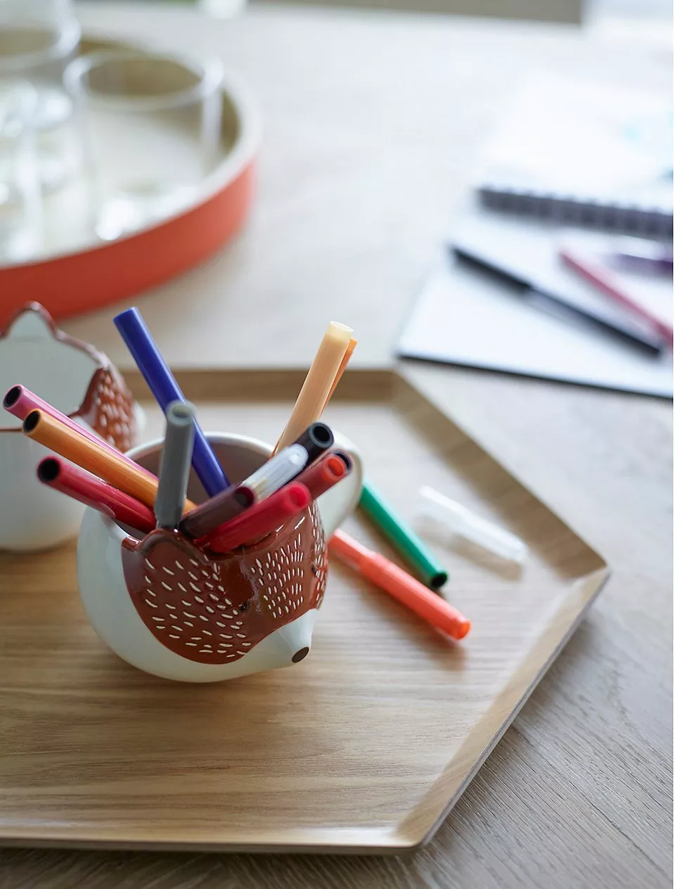 Hedgehog pencil pot holding colourful felt-tip pens on a wooden tray, with notebook and pen in background