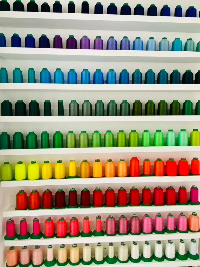 Colourful sewing thread spools arranged on white shelves in a textile studio, organised in a rainbow gradient.