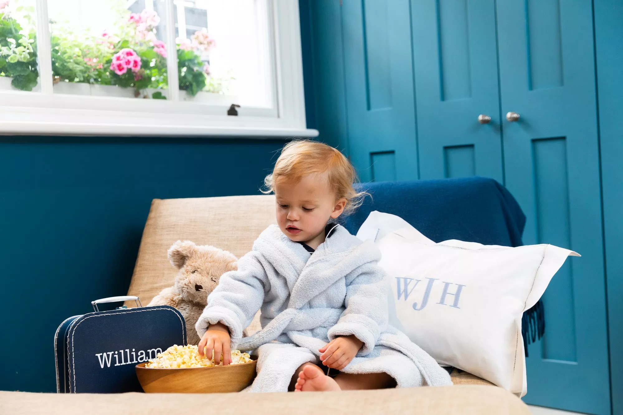Toddler in a soft bathrobe eating popcorn on a sofa, with teddy bear and blue interior décor in background