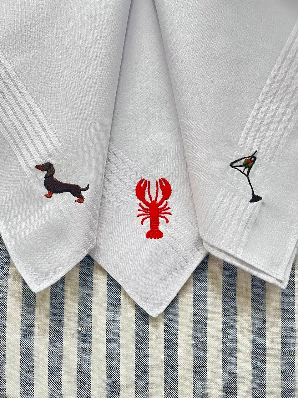 White cotton napkins with embroidered dachshund, red lobster and martini glass on blue striped linen tablecloth