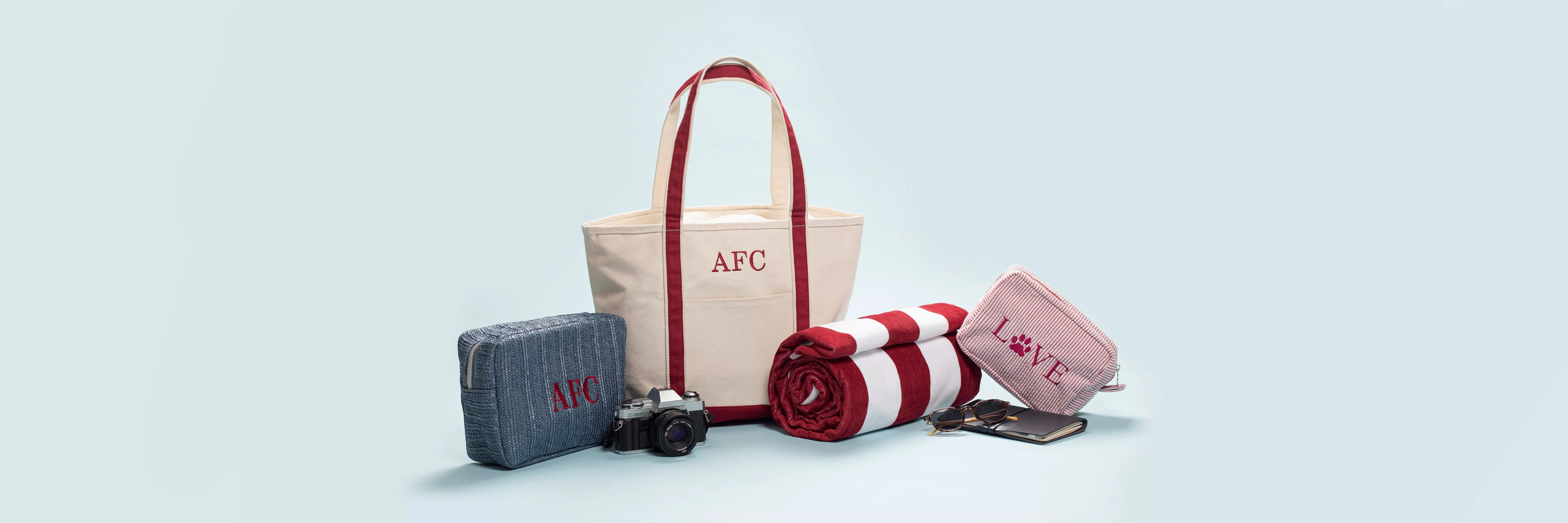 Canvas tote bag with red straps, travel pouch, striped beach towel, camera and accessories on pale blue background