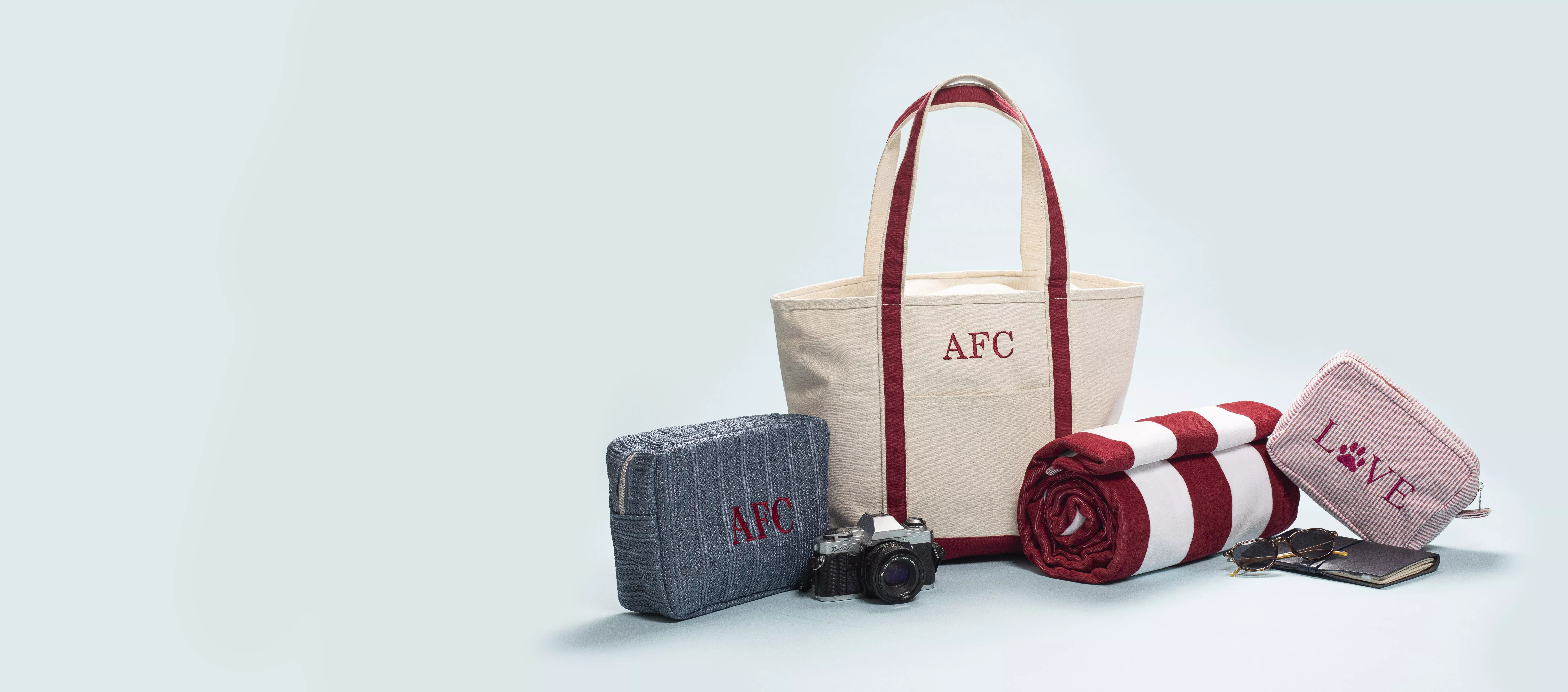 Canvas tote bag with red straps, travel pouch, striped beach towel, camera and accessories on pale blue background