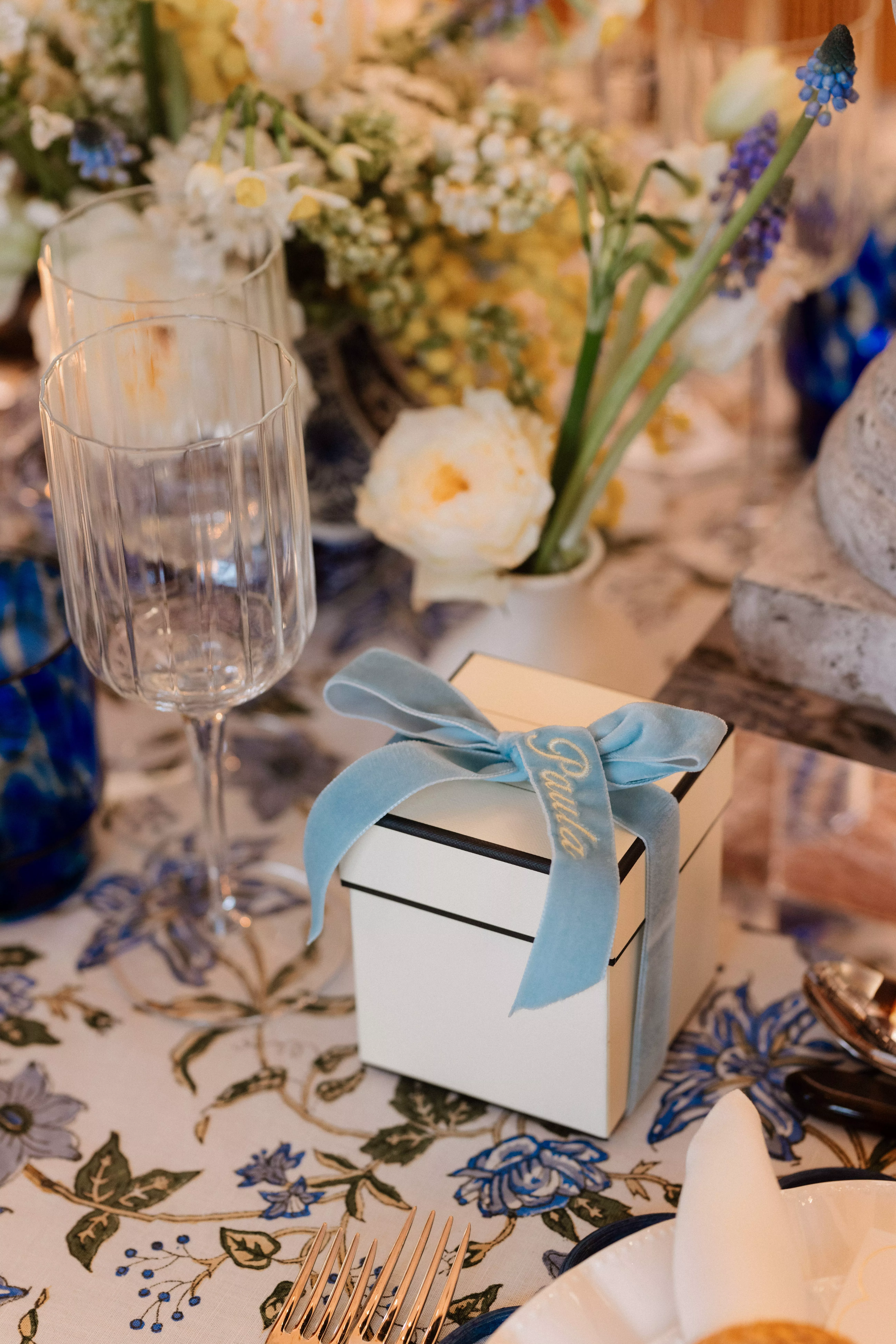 Elegant wedding table setting with floral linen, wine glass and personalised favour box tied with blue ribbon
