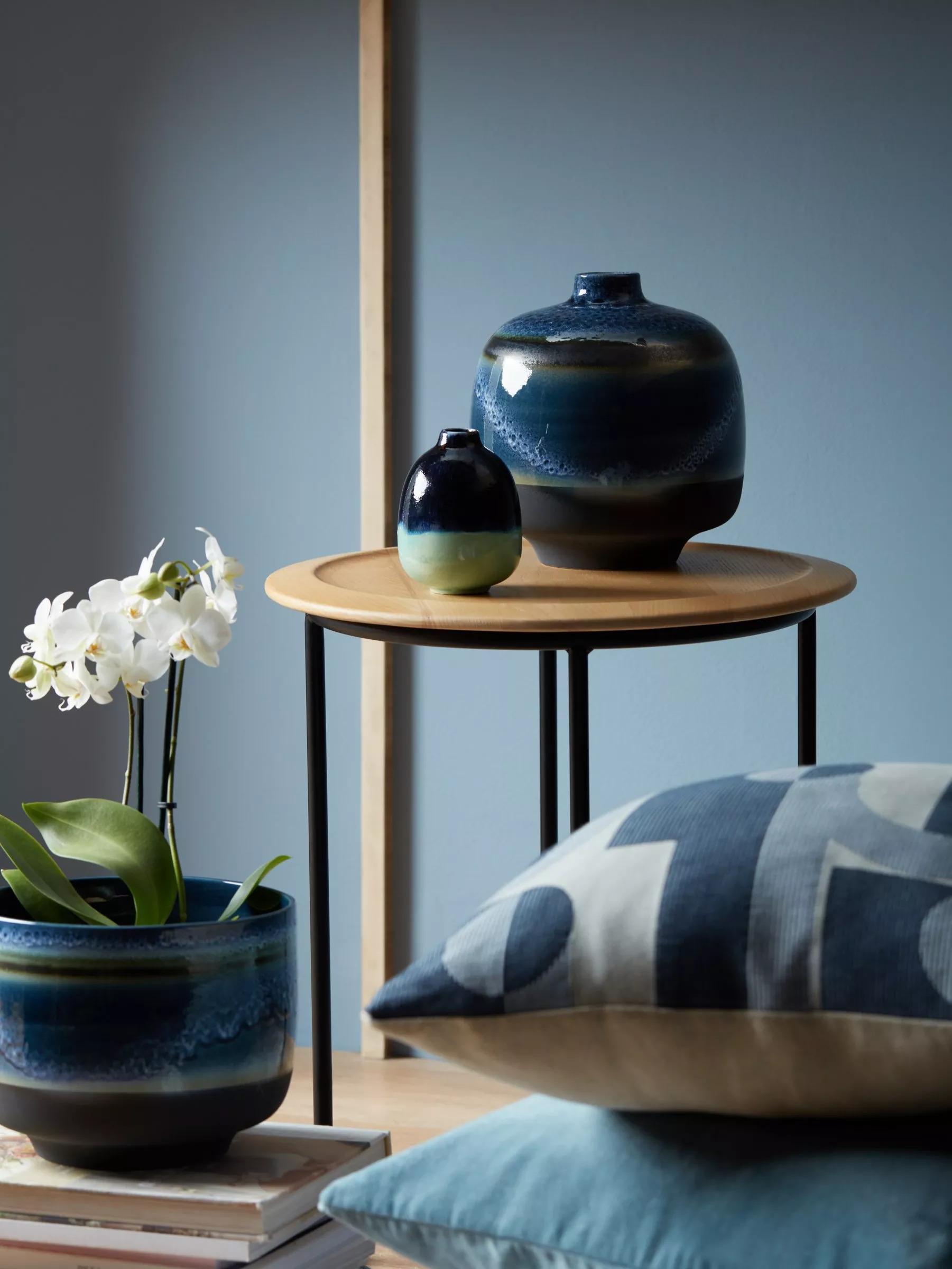 Modern home decor still life with blue glazed ceramic vases, orchid plant and cushions by a round side table
