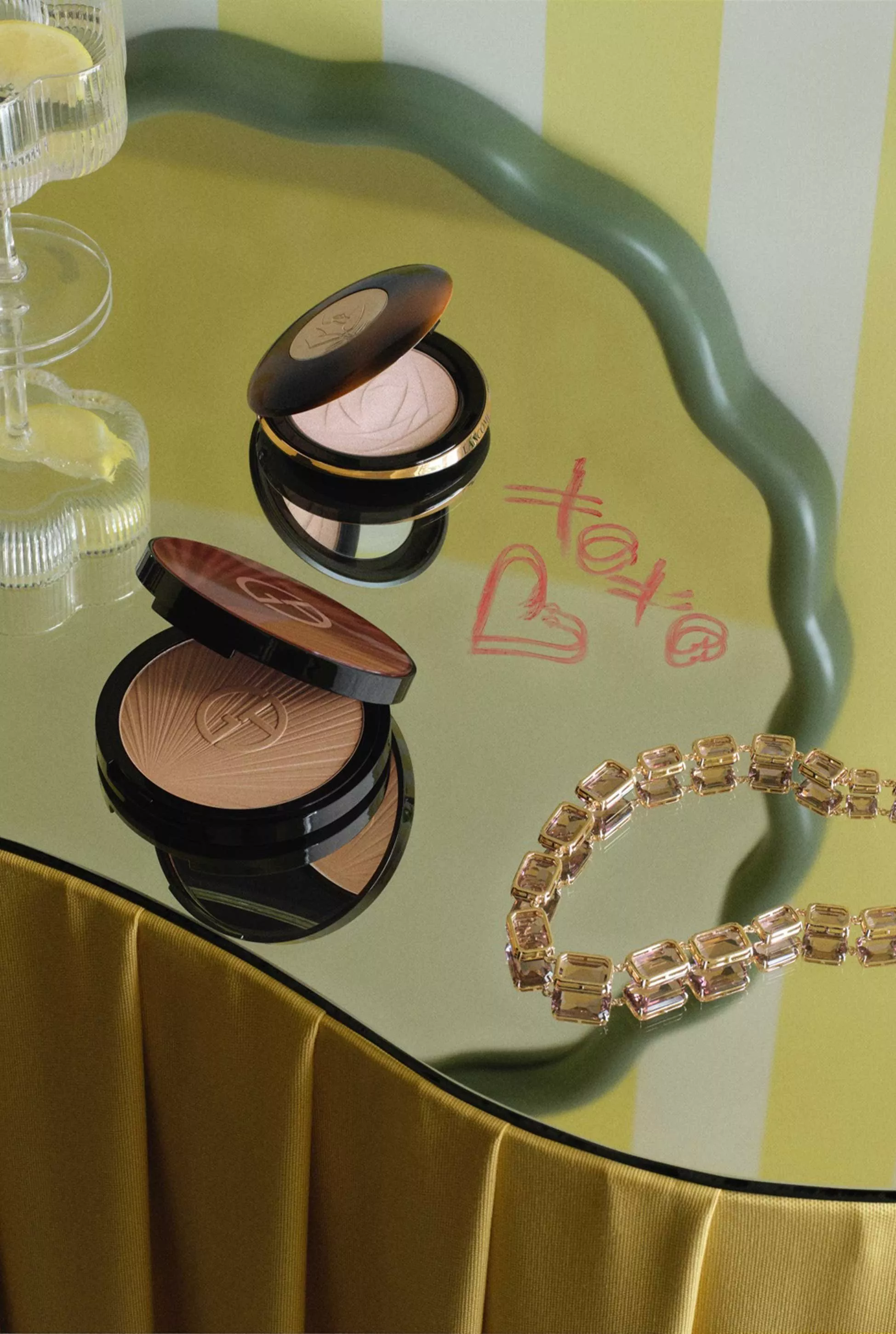Makeup compacts on a mirrored table with lipstick writing, beside a crystal necklace and fluted glasses.