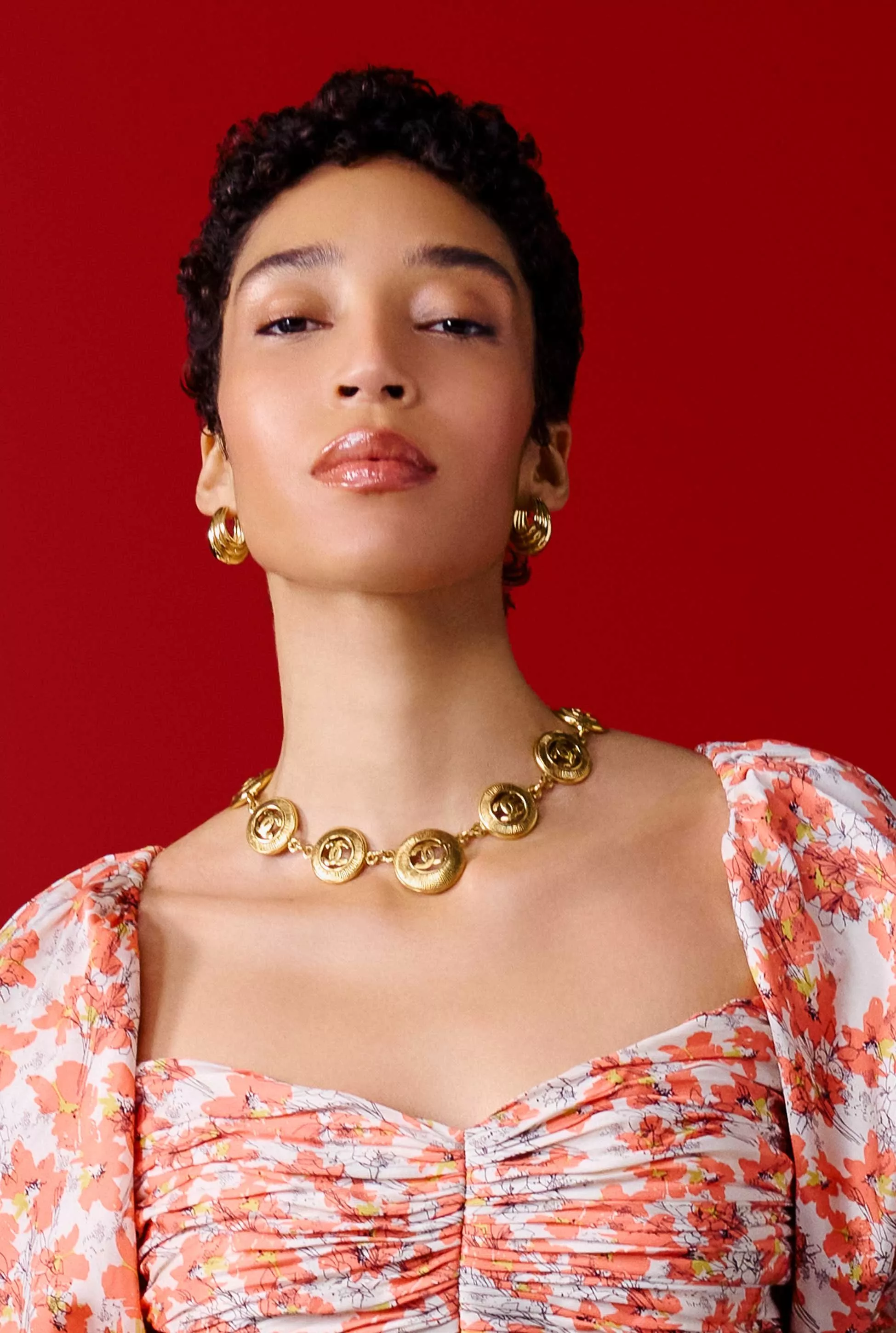A person wearing a floral dress with puffed sleeves and a gold necklace against a red background.