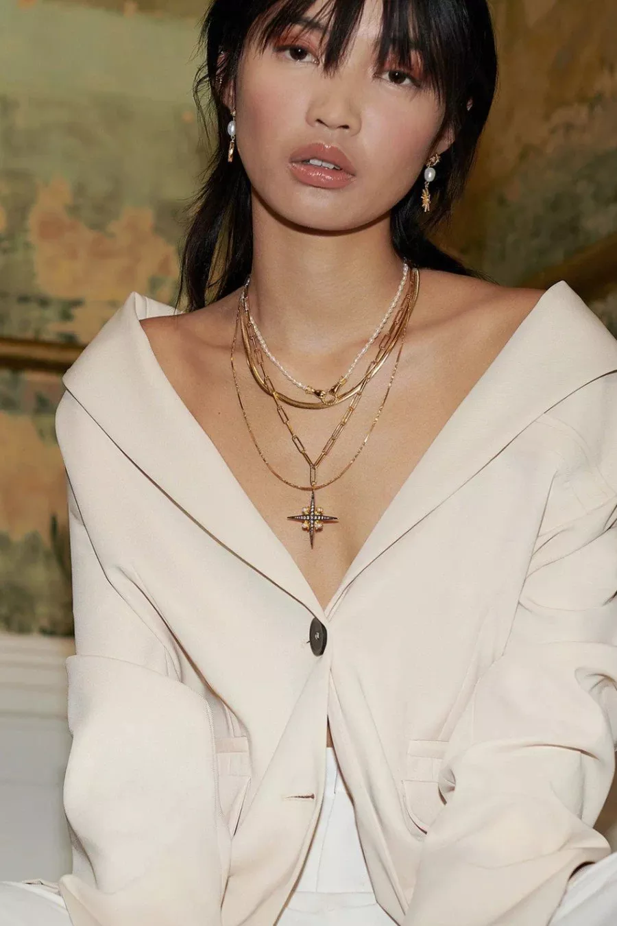 Layered gold necklaces with pearl strand and starburst pendant on woman wearing a cream blazer, close-up jewellery shot