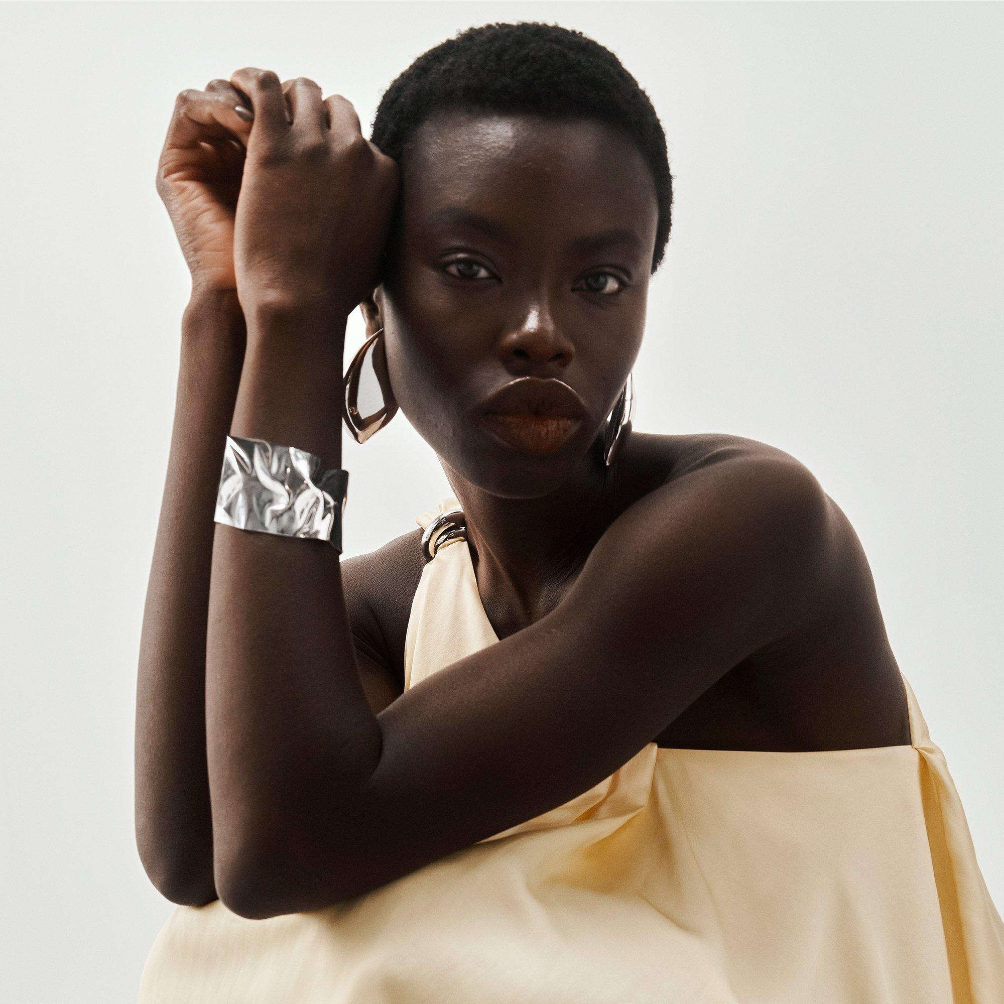 Woman in a cream dress poses with silver cuff bracelet and large hoop earrings.