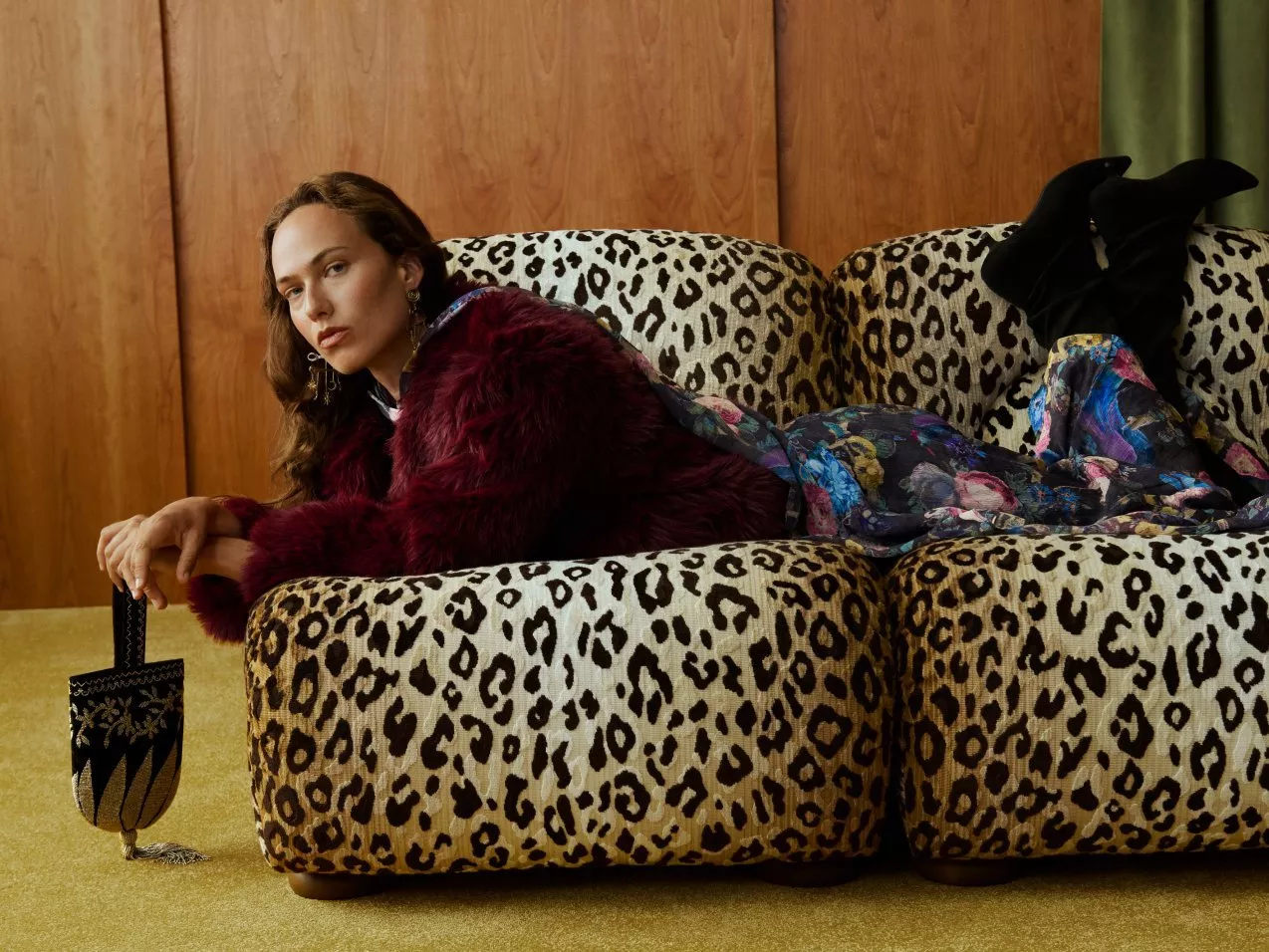 Fashion portrait of woman in burgundy faux fur and floral dress on leopard print sofa, holding clutch bag