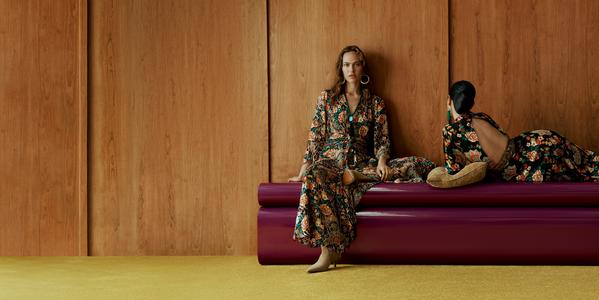 Two women modelling floral maxi dresses on a purple sofa against a wood-panel wall, modern fashion editorial.
