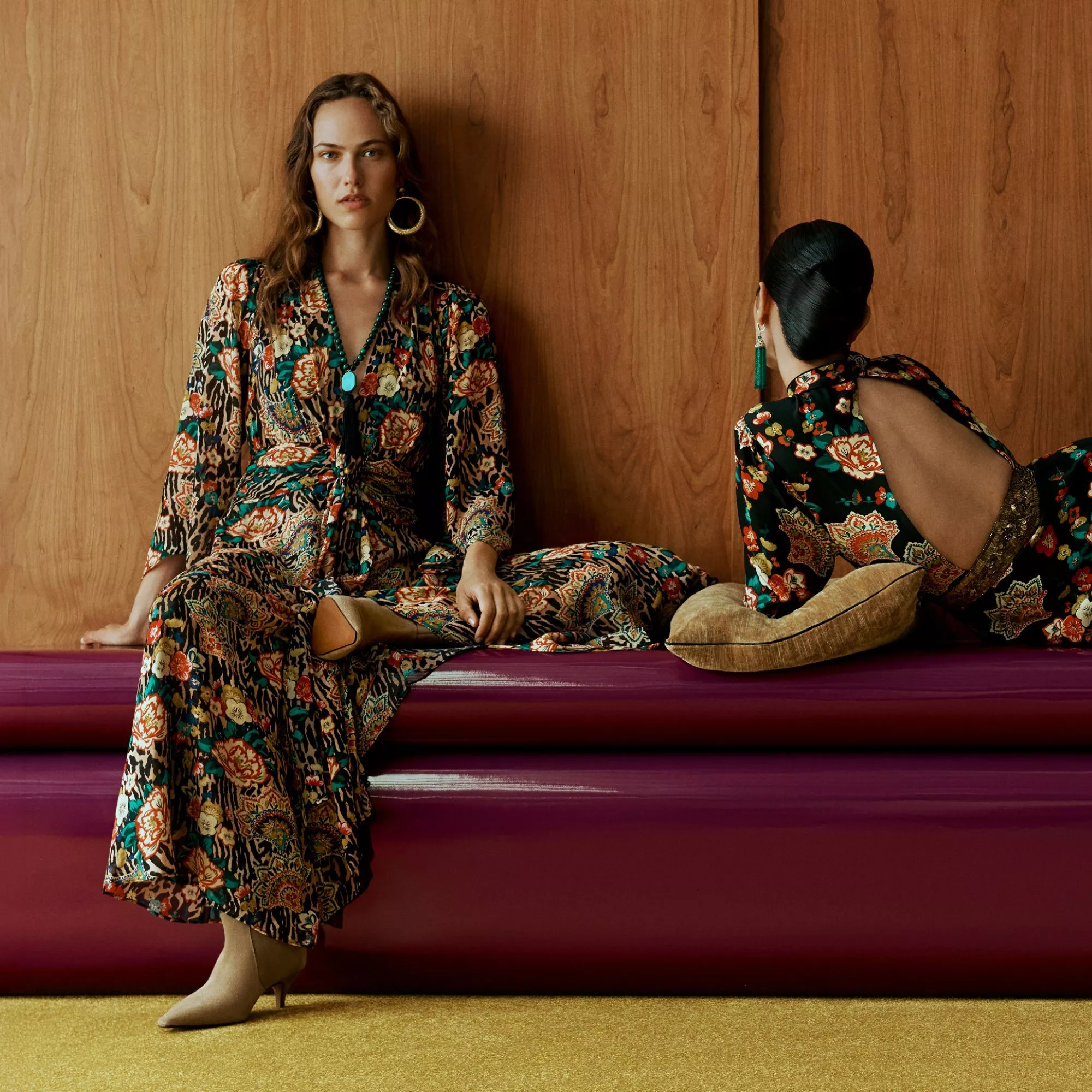 Two women in floral maxi dresses posing on a plum sofa against a wooden wall, modern fashion editorial.