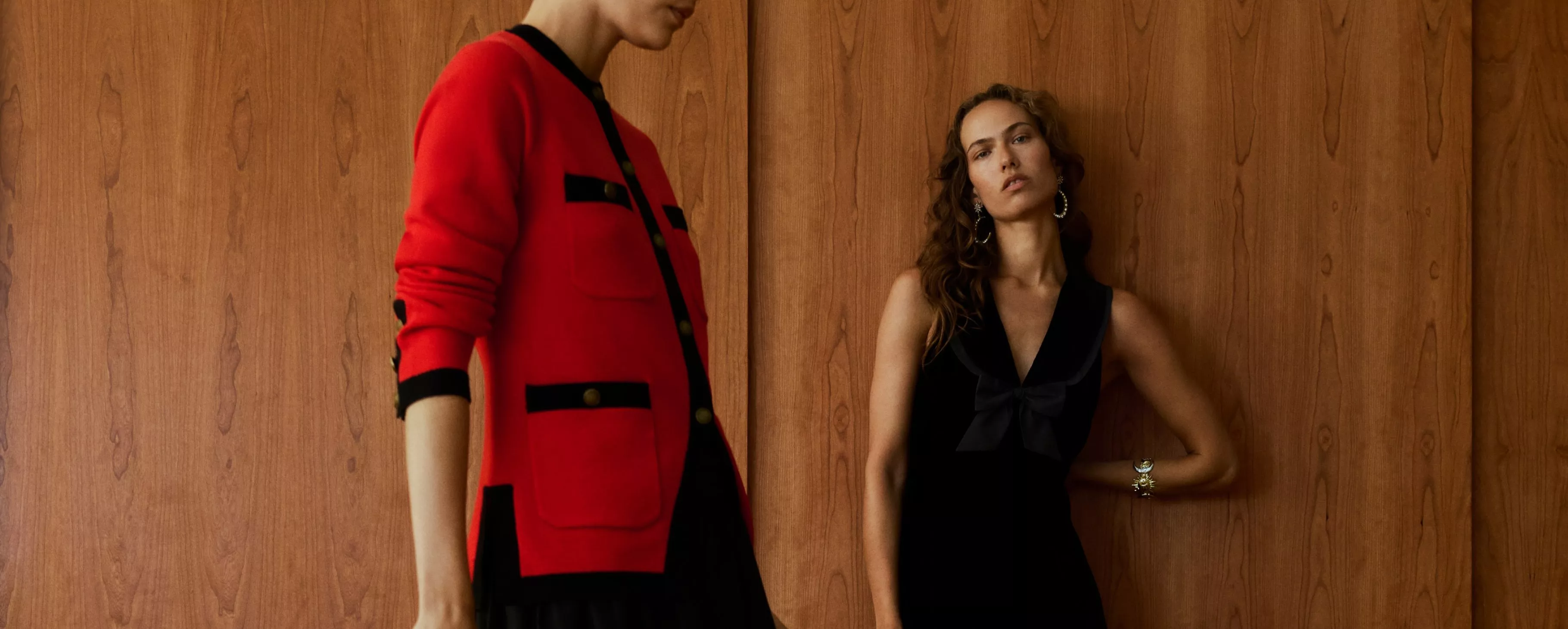 Fashion models posing by a wooden wall, one in a red cardigan and one in a black dress with earrings