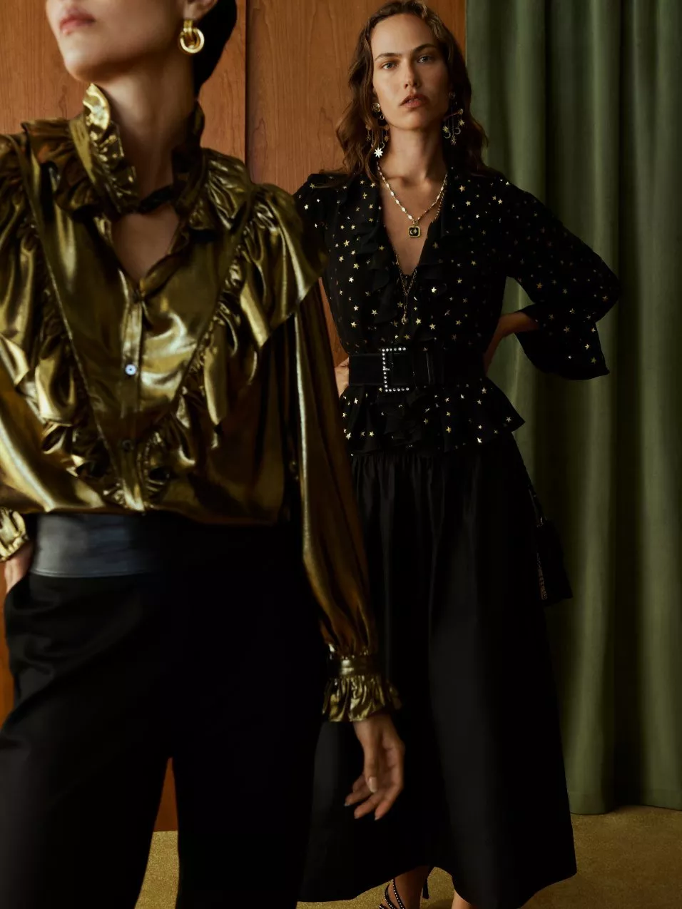 Two women, one in a gold satin blouse and black trousers, the other in a black star print top with belt and black skirt