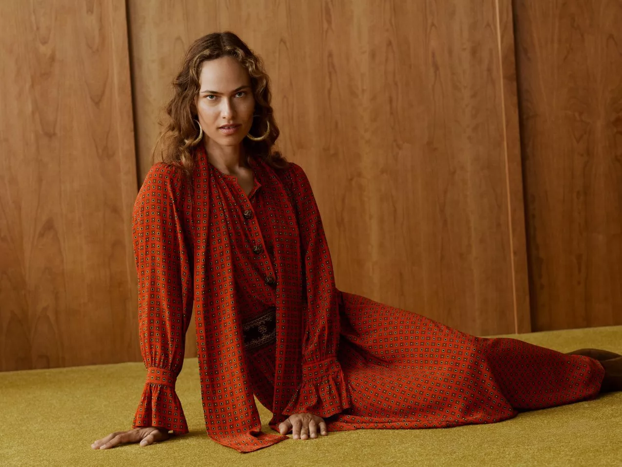 Woman in red patterned maxi dress posing on carpet floor, against wood panelling.