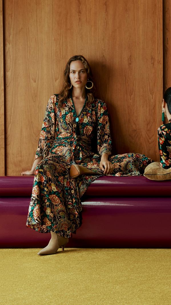Two women modelling floral maxi dresses on a purple sofa against a wood-panel wall, modern fashion editorial.