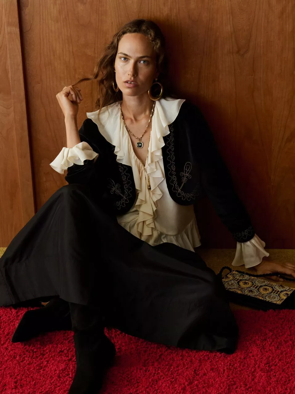 Fashion portrait of woman in black embroidered jacket and ruffled blouse, seated on red carpet by wood panel wall