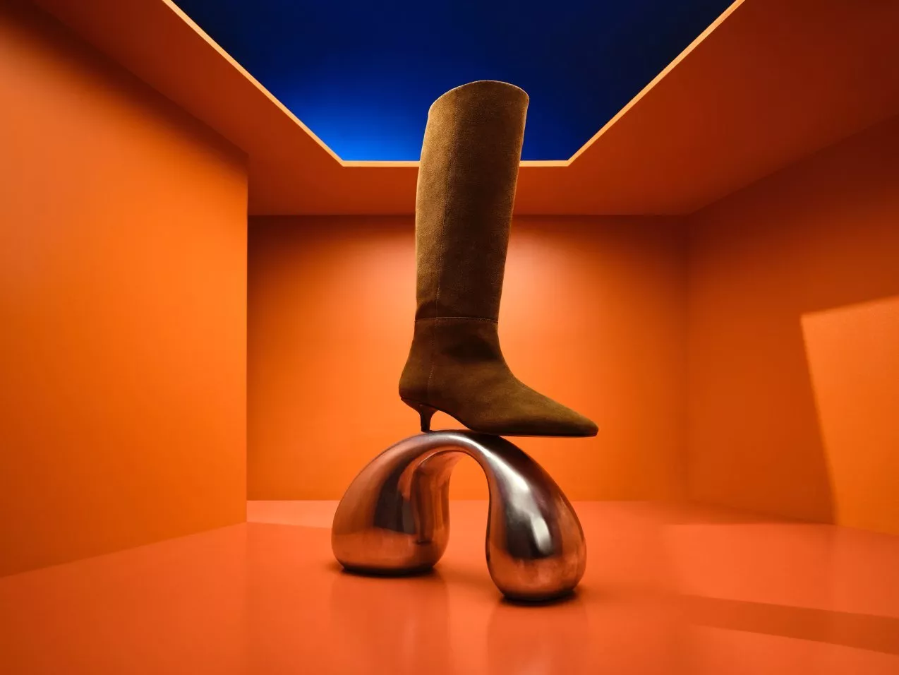 Brown suede knee-high boot on mirrored chrome sculpture in an orange room with blue ceiling