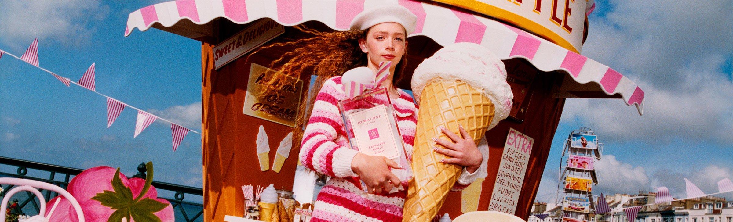 A woman in a pink-striped outfit and white beret holds an oversized ice cream cone and a large Jo Malone perfume bottle in front of a whimsical ice cream stand with pink-and-white striped decor, set against a bright, festive fairground backdrop.