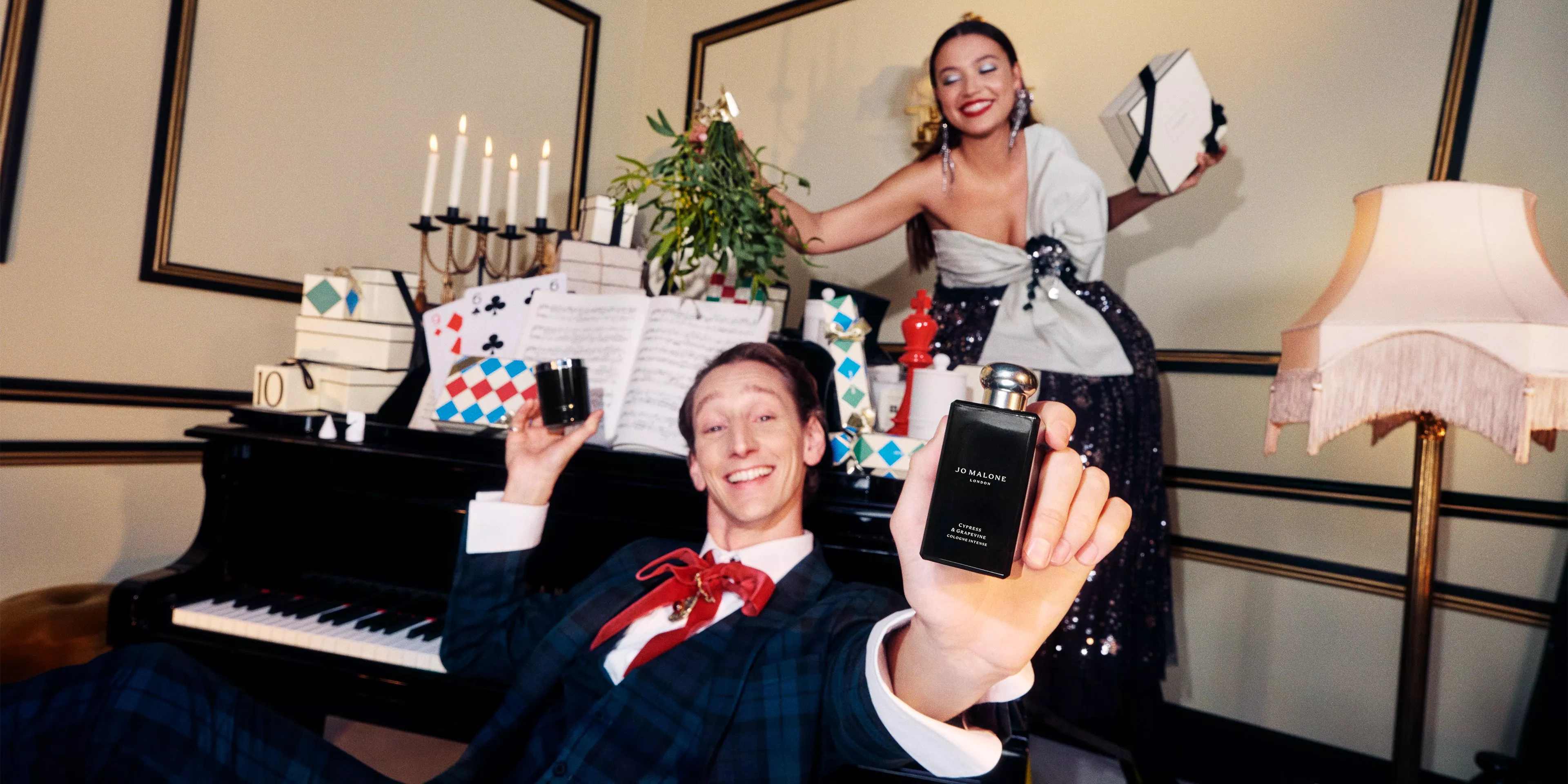 man and woman in smart suit and dress around a piano holding jo malone london products