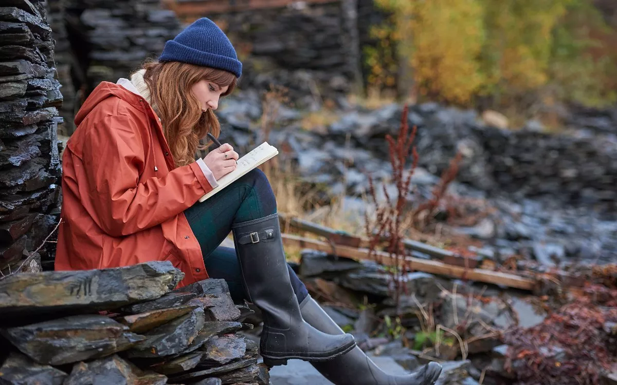 Woman in red raincoat and beanie journalling outdoors on rocky ruins, autumn landscape travel inspiration